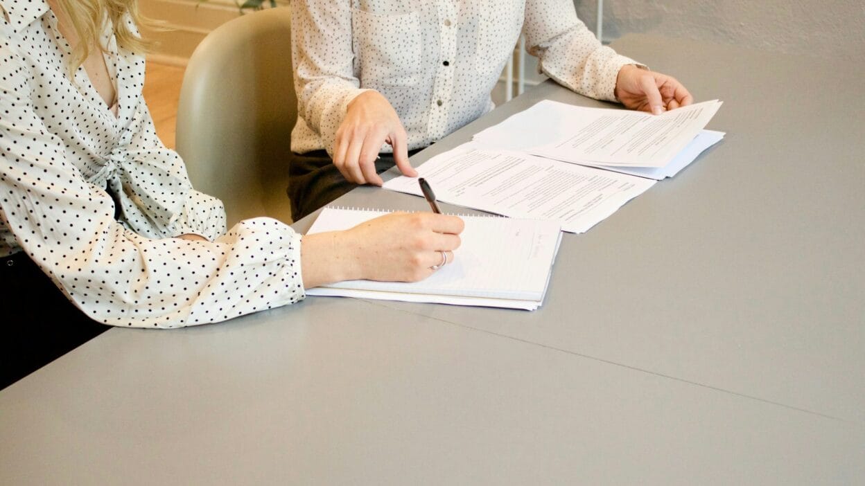 Dwie kobiety analizujące dokumenty finansowe przy biurku, prowadzące dyskusję i sporządzające notatki.