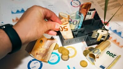 Osoba wkładająca banknot 50 euro do czarnej kasetki na pieniądze, otoczonej monetami euro, dokumentami finansowymi i kluczami do nieruchomości.
