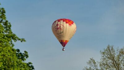 Kolorowy balon unoszący się na tle błękitnego nieba, otoczony zielonymi koronami drzew.